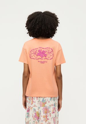 Person med krøllet hår iført en ferskenfarvet T-shirt med pink hibiscusblomstermotiv og Hawaii-øernes navne på ryggen, kombineret med et blomsterprintet skørt.