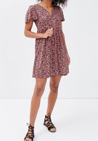 Femme portant une robe fleurie rouge à manches courtes et des sandales noires à lacets, se tenant devant un fond blanc uni.