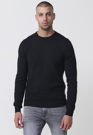 Suéter negro de punto texturizado con mangas largas y escote redondo, que presenta puños y dobladillo acanalados. Combinado con jeans grises.