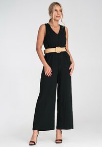 Czarny kombinezon bez rękawów z dekoltem w serek, szerokimi nogawkami, teksturowanym materiałem i brązowym, plecionym paskiem w talii. Czarne szpilki dopełniają całość.