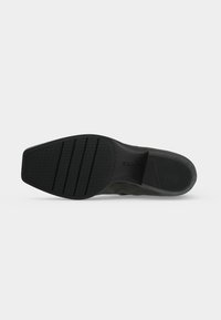 Semelle d'une chaussure habillée noire avec une semelle en caoutchouc texturée et un talon légèrement surélevé, positionnée sur un fond uni gris clair.