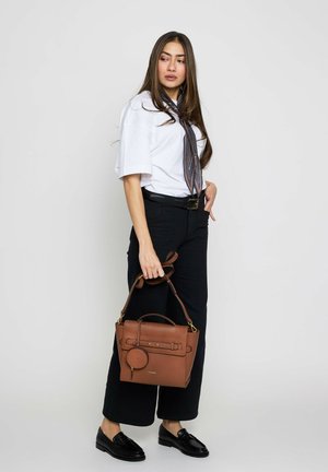 Vrouw draagt een wit overhemd, zwarte broek, patroon halsdoek, houdt een bruine leren handtas vast, staat tegen een effen achtergrond, kijkt opzij.