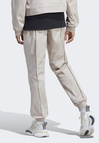 Beige träningsoveraller med elastiska muddar, sömmar på sidan och diskret logotyp. Kombineras med vita sneakers med svarta ränder och strukturerad sula.