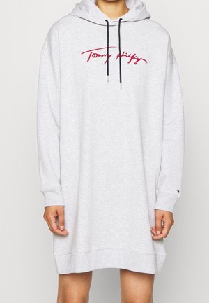 Jasnoszara oversize sukienka z kapturem z czarnymi ściągaczami, czerwone logo w stylu podpisu na piersi, noszona przez osobę z widocznymi rękami i nogami.