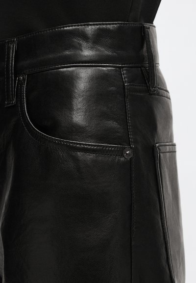 Black leather pants showing side pocket, belt loops, and seam details against black clothing and white background.
