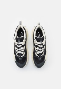 Ténis atléticos preto e branco com parte superior em malha, bico arredondado, sola de borracha grossa e detalhes contrastantes. Apresenta atacadores e uma aba de puxar na parte traseira.