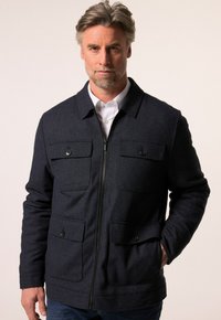 Námořnická modrá bunda vyrobená z texturované tkaniny, s předním zipem, dvěma náprsními kapsami a dvěma dolními předními kapsami se zapínáním na knoflíky.