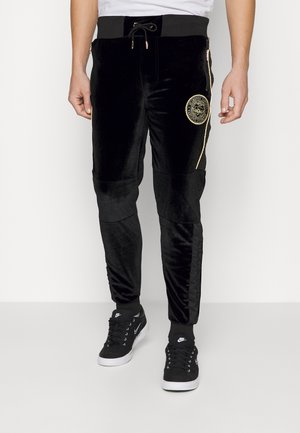 Mann trägt schwarze Velours-Jogginghose mit goldenem Besatz und Emblem, kombiniert mit schwarzen Nike-Sneakern und schwarzen Socken.
