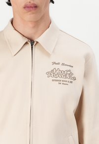 Jachetă bej cu închidere cu fermoar și guler. Prezintă broderie în culoare maro, cu textul "Full Service Huf's Paradise." Textură netedă.