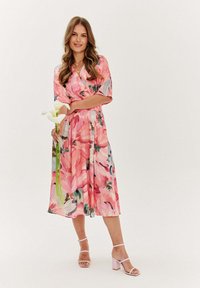 Rúžový kvetinový midi šaty s krátkymi rukávmi, s viazaním a splývavou sukňou. Modelka drží biele ľalie a má na sebe ružové sandále na blokovom podpätku.