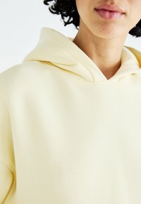 Yellow cotton hoodie with a soft texture, large front pocket, and a drawstring hood. Overlapping fabric at the neckline, minimal stitching visible.