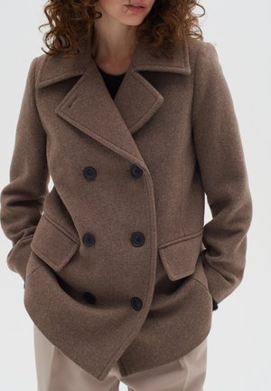 Femme portant un manteau en laine marron à double boutonnage avec de grands revers et les mains dans les poches, assorti à un pantalon beige clair.