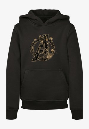 Felpa nera con logo dorato degli Avengers, abbinata a leggings verde oliva con pannelli trasparenti. Modella indossa scarpe sportive bianche.