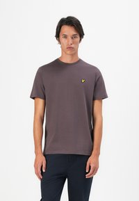 Junger Mann trägt ein dunkelgraues T-Shirt mit kurzen Ärmeln und einem kleinen gelben Vogel-Logo auf der linken Brust, steht vor einem schlichten weißen Hintergrund.