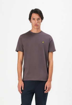 Jeune homme portant un t-shirt gris foncé à manches courtes avec un petit logo d'oiseau jaune sur la poitrine gauche, se tenant devant un fond blanc uni.