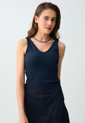 Jonge vrouw met lang bruin haar, gekleed in een marineblauw mouwloos topje, bijpassende rok en een blauwe kralenketting, staand tegen een effen achtergrond.