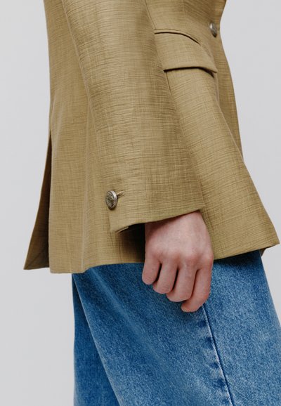 Χέρι που ακουμπάει στο πλάι, φοράει ταγιέρ καφέ ανοιχτού χρώματος με υφή και λεπτομέρειες από κουμπιά, καθώς και μπλε τζιν, πάνω σε απλό φόντο.