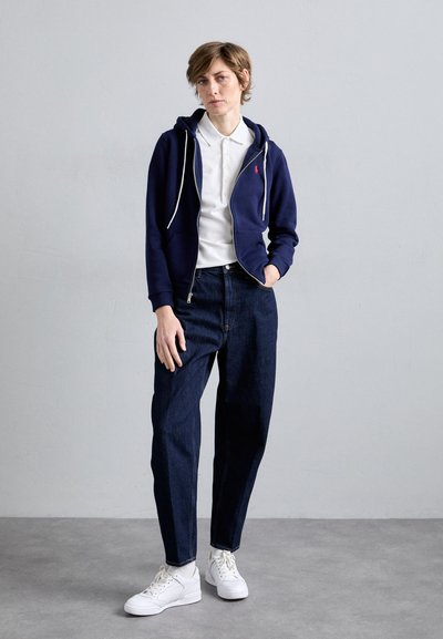 Marinefarbener Reißverschluss-Hoodie mit weißen Kordeln über einem weißen Poloshirt, kombiniert mit dunkelblauen Jeans und weißen Sneakers, vor einem neutralen Hintergrund stehend.