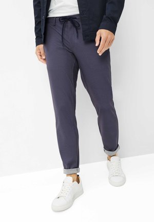 Mand iført mørkeblå bukser med snøre, der er rullet op, hvide sneakers, hvid skjorte og mørk jakke, stående mod en ensfarvet baggrund.