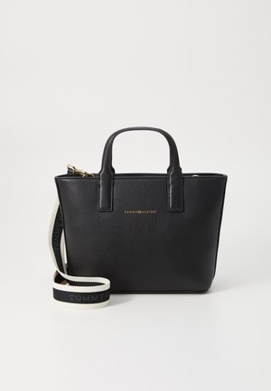 Sac à main en cuir noir avec un logo métallique doré. Il dispose de deux poignées supérieures et d'une bandoulière amovible avec un branding noir et blanc. Texture lisse.