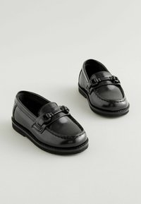 Mocasines de charol negro con un diseño cosido y acentos decorativos de hebilla. Presentan una suela de goma negra baja y una textura suave.