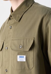 Olivgrünes Hemd mit umklappbarem Kragen, zwei Brusttaschen und einem weißen Markenetikett. Abgerundet mit schwarzen Knopfdetails und einem strukturierten Stoff.