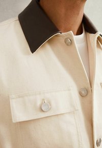 Cremefarvet jakke med krave i brunt læder, med metallic trykknapper og en brystlomme. Struktureret stof med subtile prikker.