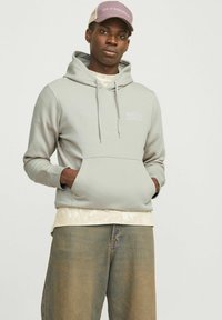Hellgrauer Hoodie mit Kängurutasche, verstellbarem Kordelzug an der Kapuze und weißem "JACK & JONES"-Logo. Getragen über einem beigen Shirt und lockeren Jeans.