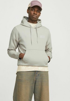 Jasnoszara bluza z kapturem i kangurzą kieszenią, regulowanym sznurkiem w kapturze oraz białym logiem "JACK & JONES". Nosić na beżowej koszulce i luźnych dżinsach.