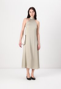 Maxi dress senza maniche di un beige chiaro con scollatura alta e design fluido. La modella tiene una piccola borsa bianca e indossa delle flats nere.
