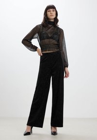 Top lungo trasparente nero con maniche lunghe sovrapposto a un bralette nero, abbinato a pantaloni neri a gamba larga a vita alta con dettagli scintillanti sottili.