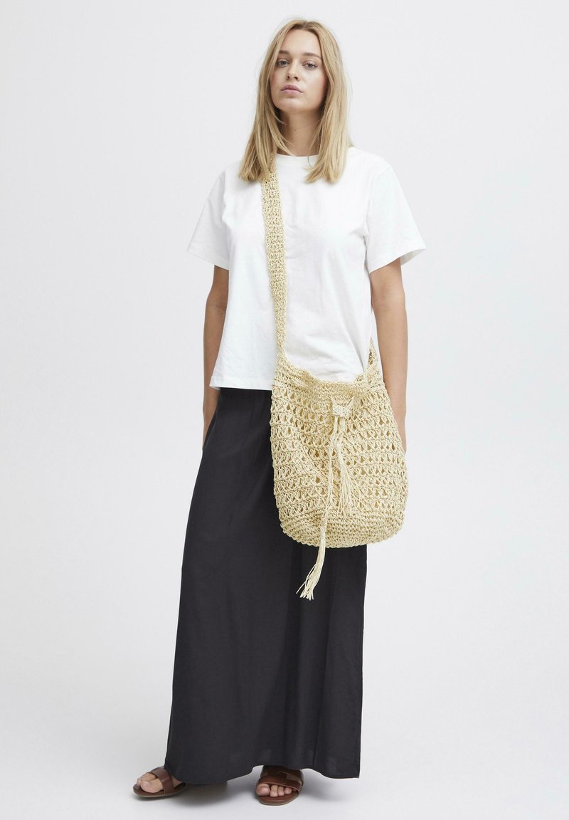 Borsa a tracolla in paglia intrecciata con chiusura a coulisse e nappina, caratterizzata da un design testurizzato a trama aperta in un colore beige chiaro.