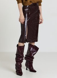Jupe en simili cuir bordeaux taille haute avec une fente frontale, portée avec des bottes bordeaux slouchy montantes jusqu'au genou et à bout pointu. Texture lisse.