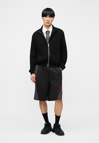 Νέος άνδρας όρθιος, φοράει μαύρο μπουφάν με φερμουάρ πάνω από λευκό πουκάμισο και γραβάτα, μαύρες και γκρίζες βερμούδες με κόκκινες λεπτομέρειες, μαύρες κάλτσες και μαύρα αθλητικά παπούτσια.