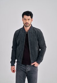 Harmaa mokkanahkainen bomber-takki, jossa on ribbineulotut hihansuut ja kaulus sekä kaksi vetoketjullista sivutaskua, puettuna tumman neuleen ja farkkujen päälle.