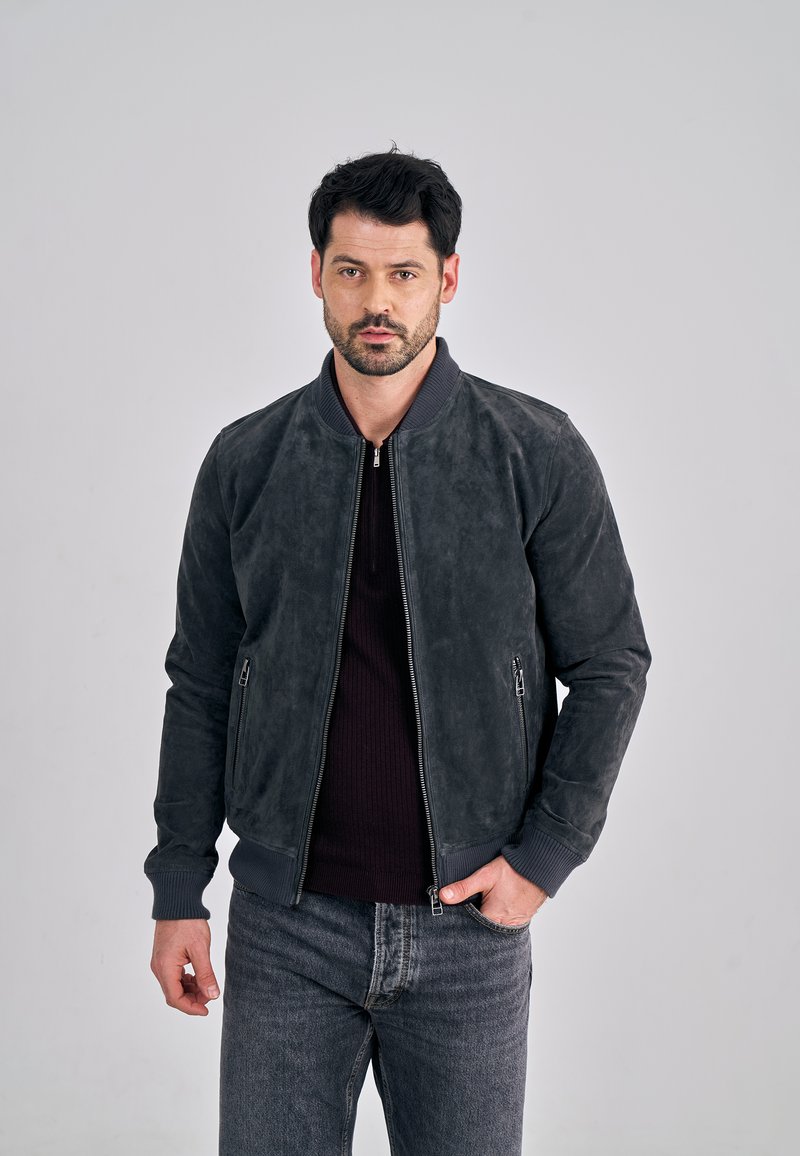 Harmaa mokkanahkainen bomber-takki, jossa on ribbineulotut hihansuut ja kaulus sekä kaksi vetoketjullista sivutaskua, puettuna tumman neuleen ja farkkujen päälle.