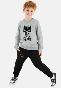 Grauer Hoodie mit schwarzem Blitzgrafik und "SEI MUTIG" Text, kombiniert mit schwarzen Jogginghosen mit Blitzakzenten. Schwarze Sneaker mit Tierdesign.
