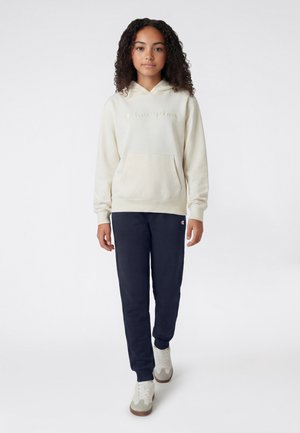 Dziewczyna z kręconymi włosami, ubrana w kremową bluzę Champion z kapturem, granatowe dresy i białe trampki, stojąca na jednolitym tle.