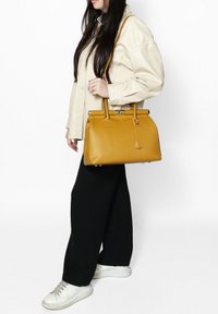 Borsa gialla strutturata con manici doppi, finitura texturizzata e chiusura a lucchetto. Abbinata a una camicia beige oversize e pantaloni neri.