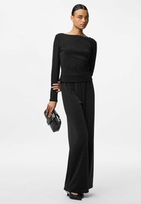 Schwarzes Langarmoberteil und weite Hose aus schimmerndem Stoff. Das Model hält eine kleine schwarze Handtasche mit silbernen Akzenten.