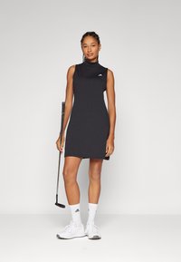 Μαύρο μίνι φόρεμα γκολφ χωρίς μανίκια με ψηλό γιακά, φέρει ένα μικρό λογότυπο. Το μοντέλο κρατάει ένα γκολφ πότερ, φορώντας λευκά παπούτσια και κάλτσες.