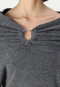 Suéter gris de punto con un escote profundo, que presenta tejido recogido alrededor del cuello y un detalle de botón decorativo en el centro.