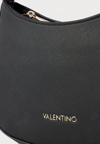 Crna teksturirana torbica s glatkom završnom obradom, s zlatnim logotipom "VALENTINO" i zatvaranjem na zatvarač na vrhu. Elegantnog i kompaktnog dizajna.