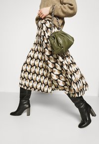 Faltenrock mit einem geometrischen Muster in Braun, Creme und Beige, kombiniert mit einer grünen Handtasche und schwarzen kniehohen Stiefeln mit Absatz.