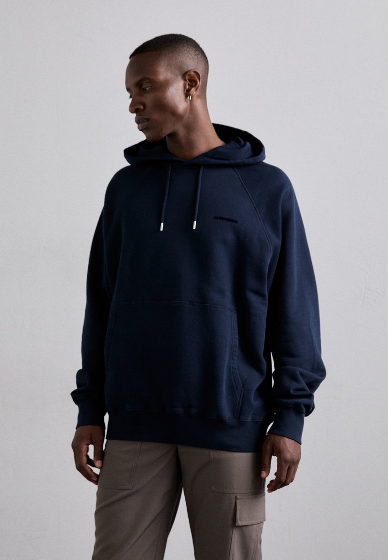 Felpa oversize blu navy con tasca frontale, coulisse regolabile e polsini a coste. Presenta un logo minimalista sul petto.