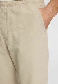 Beige texturerade byxor med elastisk midja och sidofickor, med en subtil lyster och slät tygfinish.