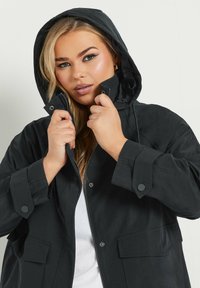 Svart huvtjacka med dragkedja och knäppning, som har två framfickor och en avslappnad passform. Slät textur och lätt material.