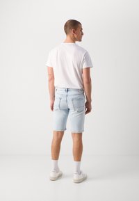 Shorts en denim bleu clair avec une coupe droite et un ourlet effiloché, associés à un t-shirt blanc. Baskets blanches avec logo visible et chaussettes.