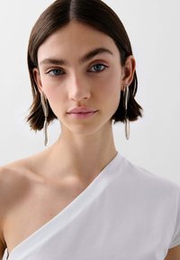Silberne Tropfenohrringe hängen von den Ohren des Models und ergänzen ein weißes, einseitig geschnittenes Oberteil mit glatter Textur und taillierter Passform.