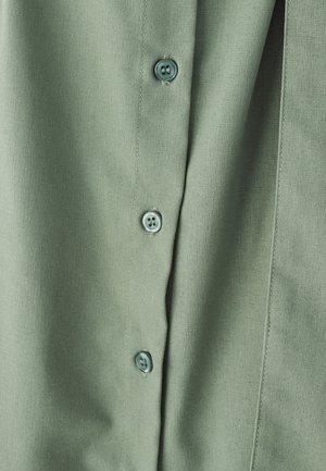 Grøn stofskjorte med en glat tekstur, knaplukning med lyseblå knapper og diskret syning langs stolpen.